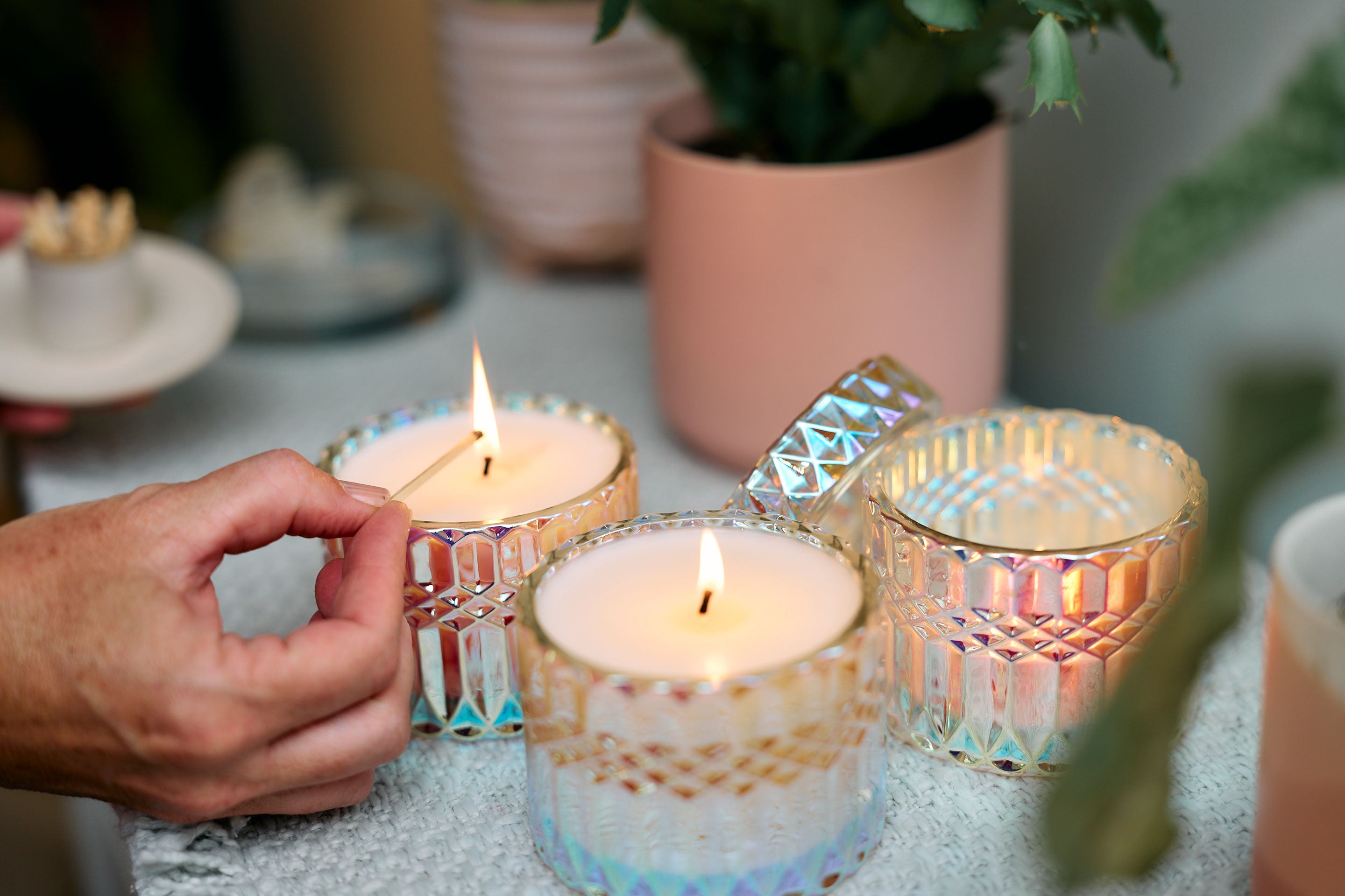 Sunbeam Candles SunCatcher scented apricot, coconut & soy wax iridescent container candles being lit by a hand on a table with plants behind them