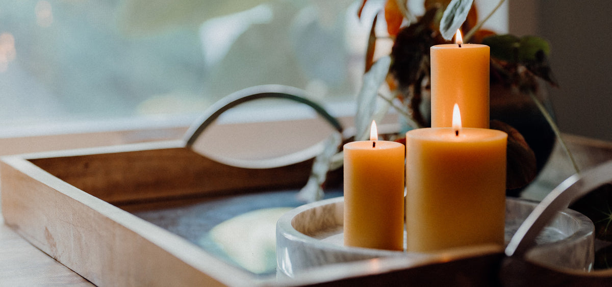 Three beeswax pillars lit on a table in front of a window with a plant in the background