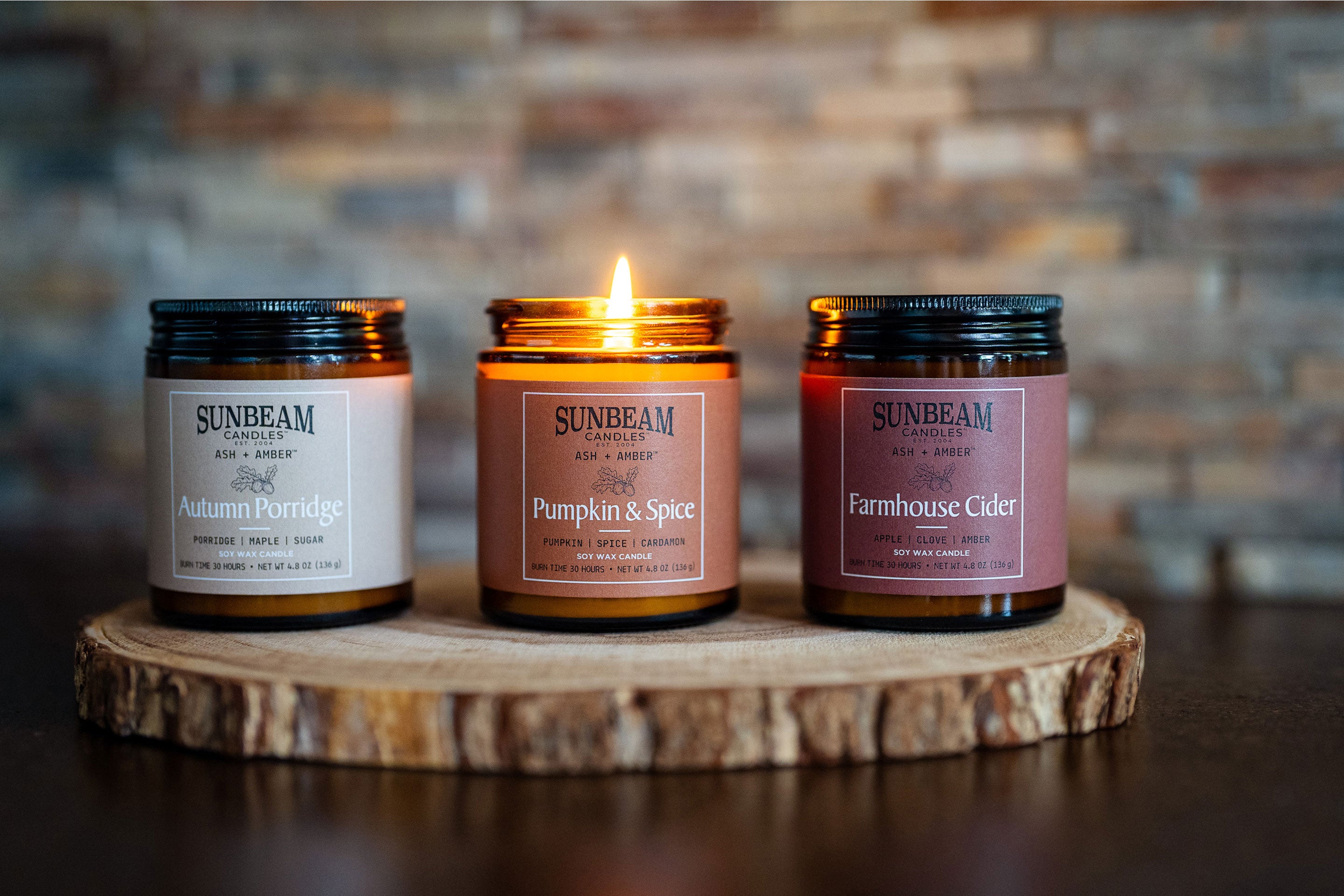 Sunbeam Candles Ash + Amber lit and unlit Fall Scent Soy Wax jar candles on a slice of a tree stump on a black table with a brick wall behind them.