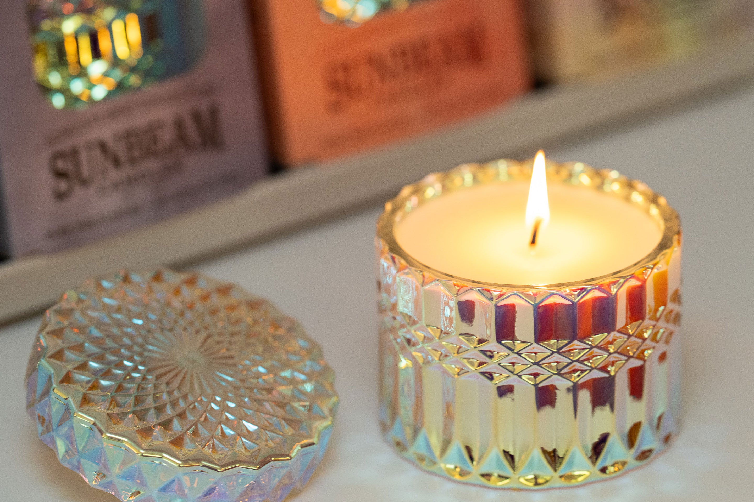 Sunbeam Candle SunCatcher scented apricot, coconut & soy wax iridescent container candlelit with lid next to it and blurred view of boxed SunCatchers behind it