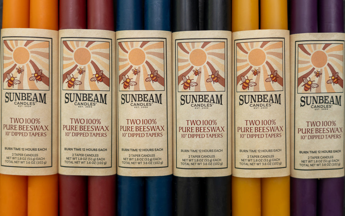 Sunbeam Candles 10" dipped beeswax tapers with wrapped labels in various colors next to each other - Apricot, Crimson, Indigo, Midnight Black, Natural Gold & Plum