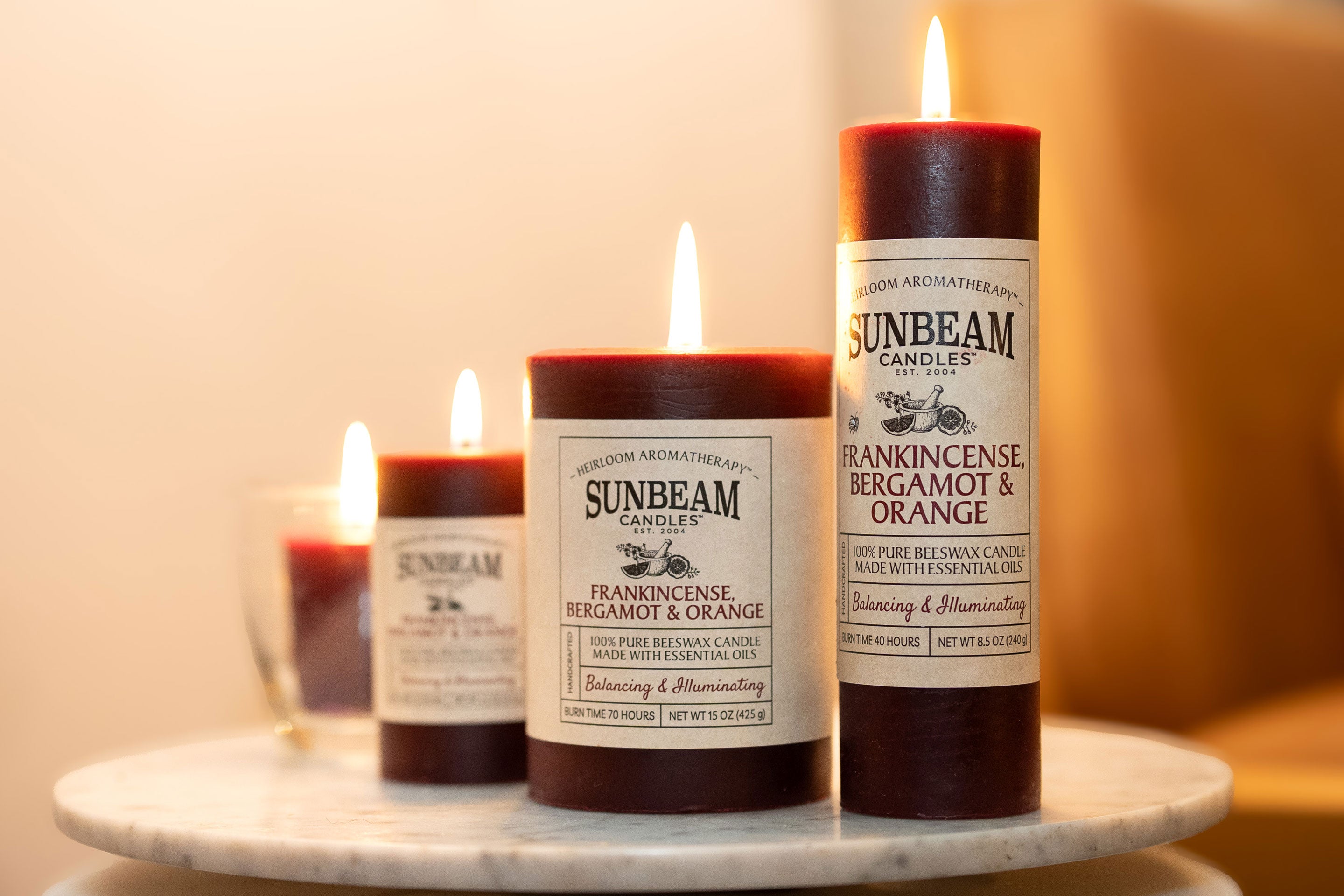 Sunbeam Candles pure burgundy Beeswax Frankincense Bergamot & Orange lit pillar candles on a marble stand with a white and yellow background