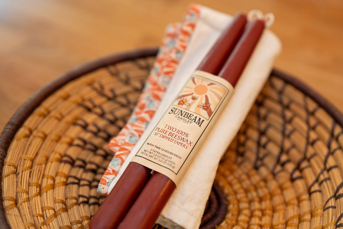 One pair of Sunbeam Candles 10" Dipped Beeswax Crimson Tapers with a label on a napkin resting on a wicker coaster.