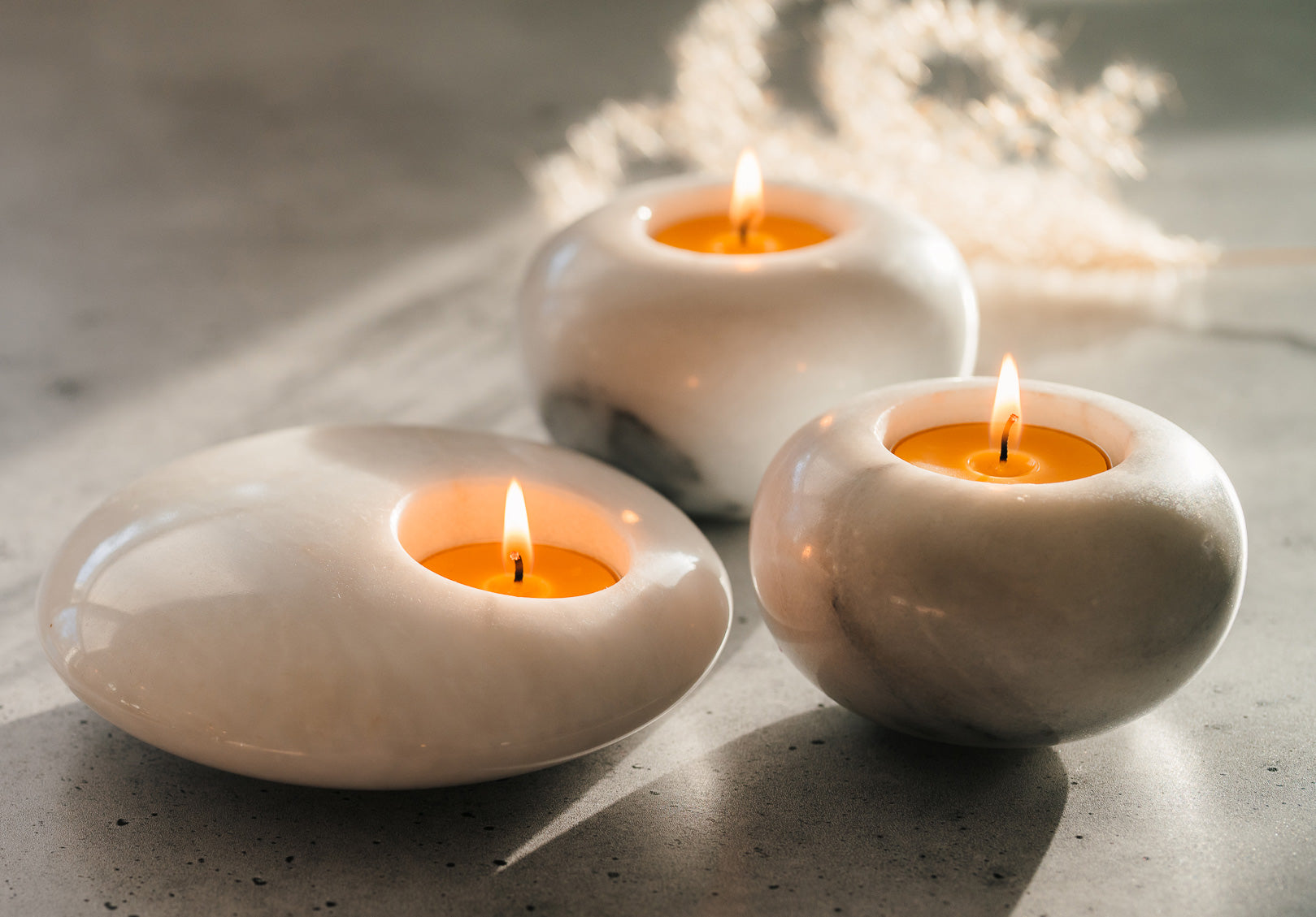 Three white ceramic candle holders with Sunbeam Candles pure beeswax tealight candles lit on a concrete surface.