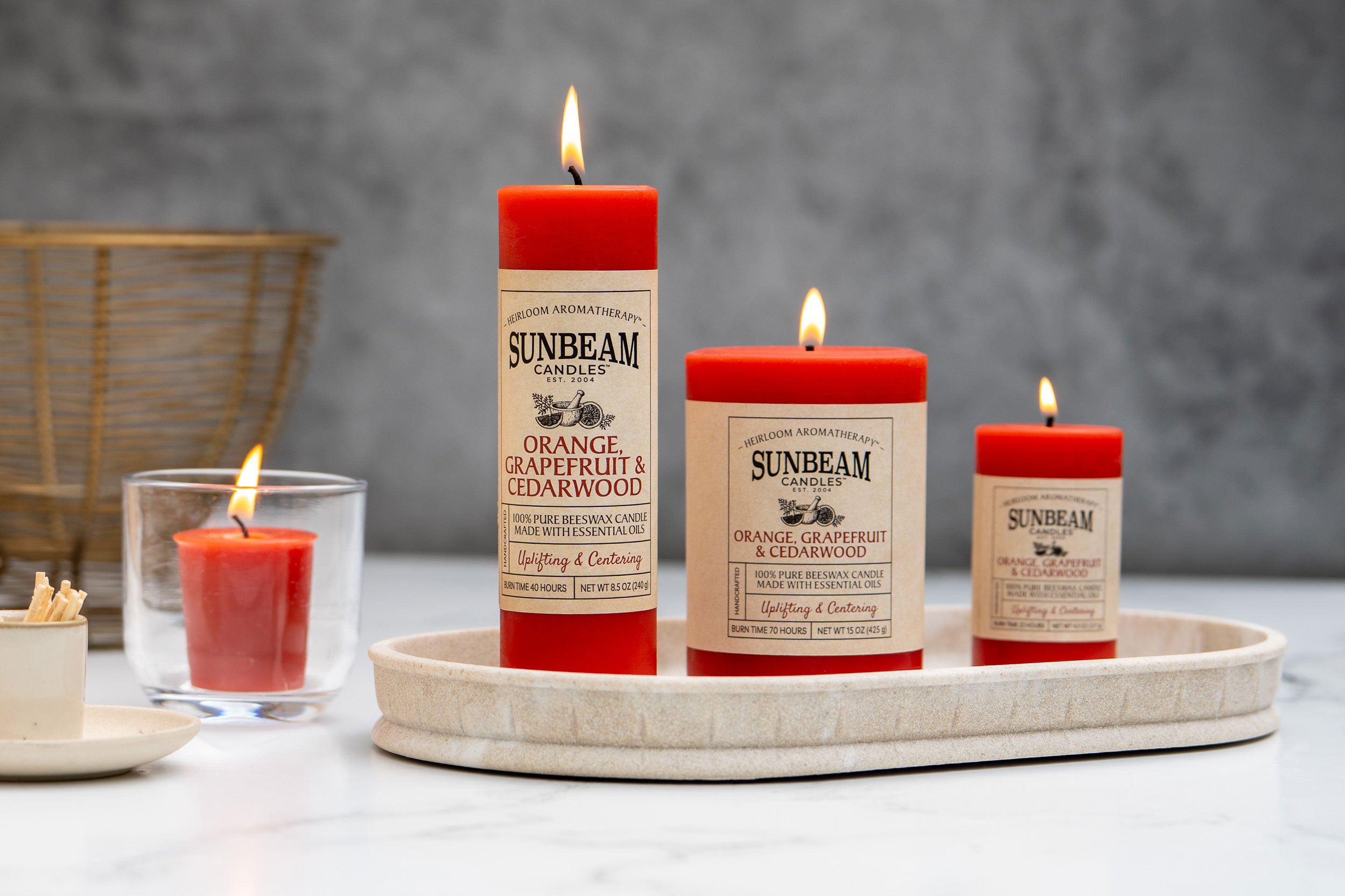 Sunbeam Candles Pure Beeswax reddish-orange Orange, Grapefruit & Cedarwood pillar & votive candles on a table with a grey background and wicker basket behind the candles