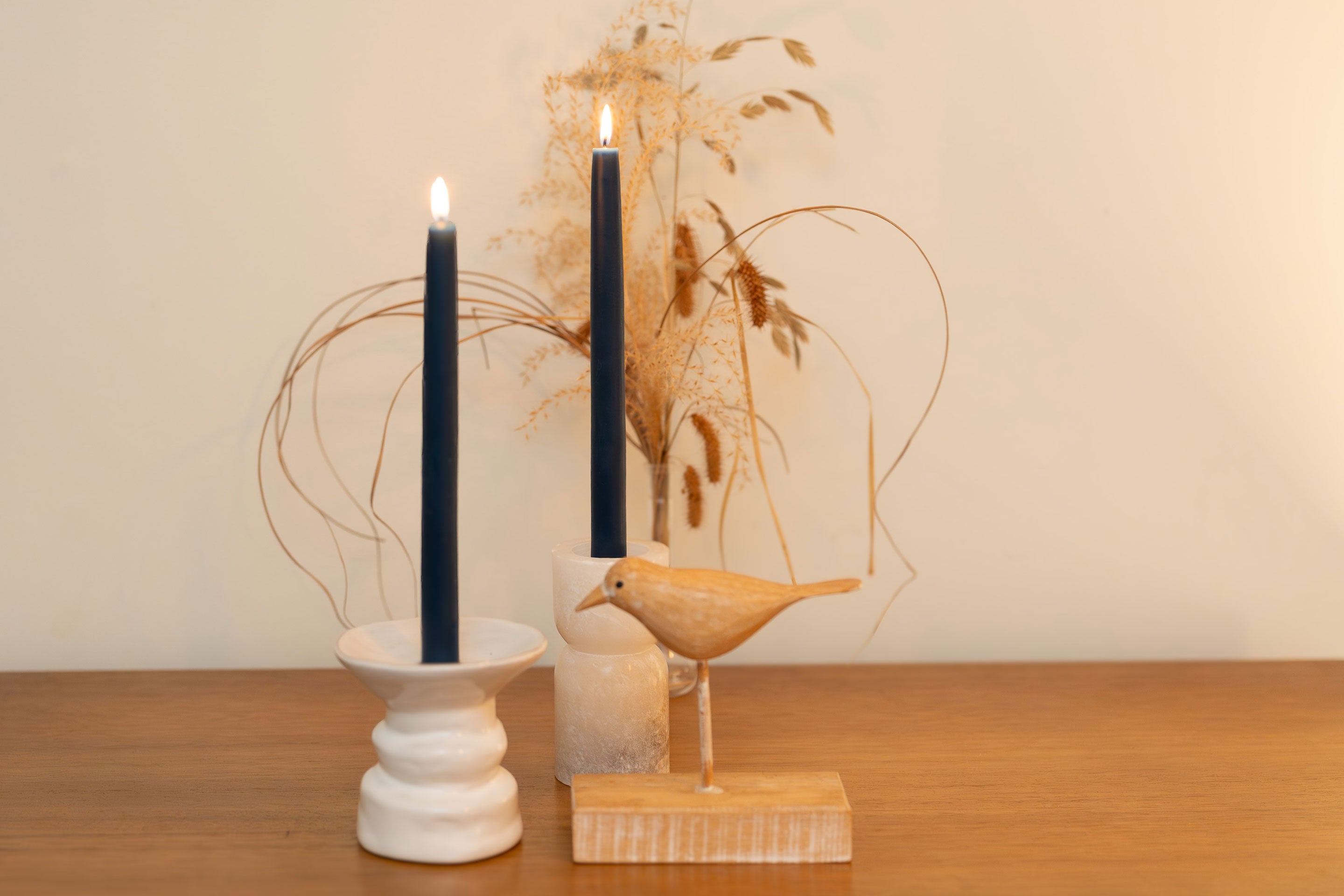 Two Sunbeam Candles 10" Dipped Beeswax Indigo Tapers lit in candle holders with a plant behind them and a bird sculpture next to them.
