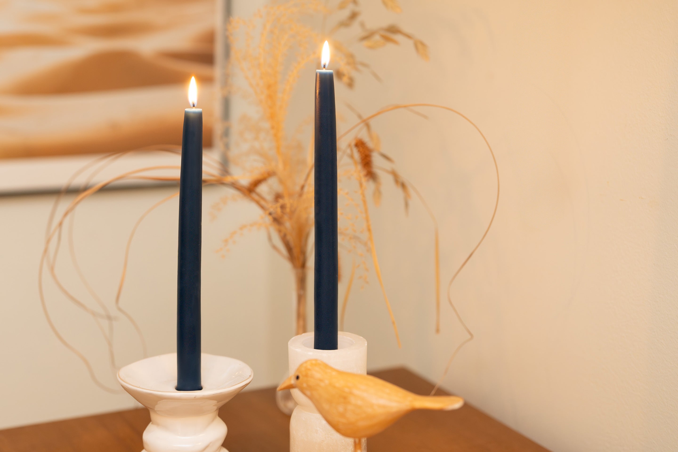 Two Sunbeam Candles 10" Dipped Beeswax Indigo Tapers lit in candle holders with a plant behind them and a cream colored wall behind.