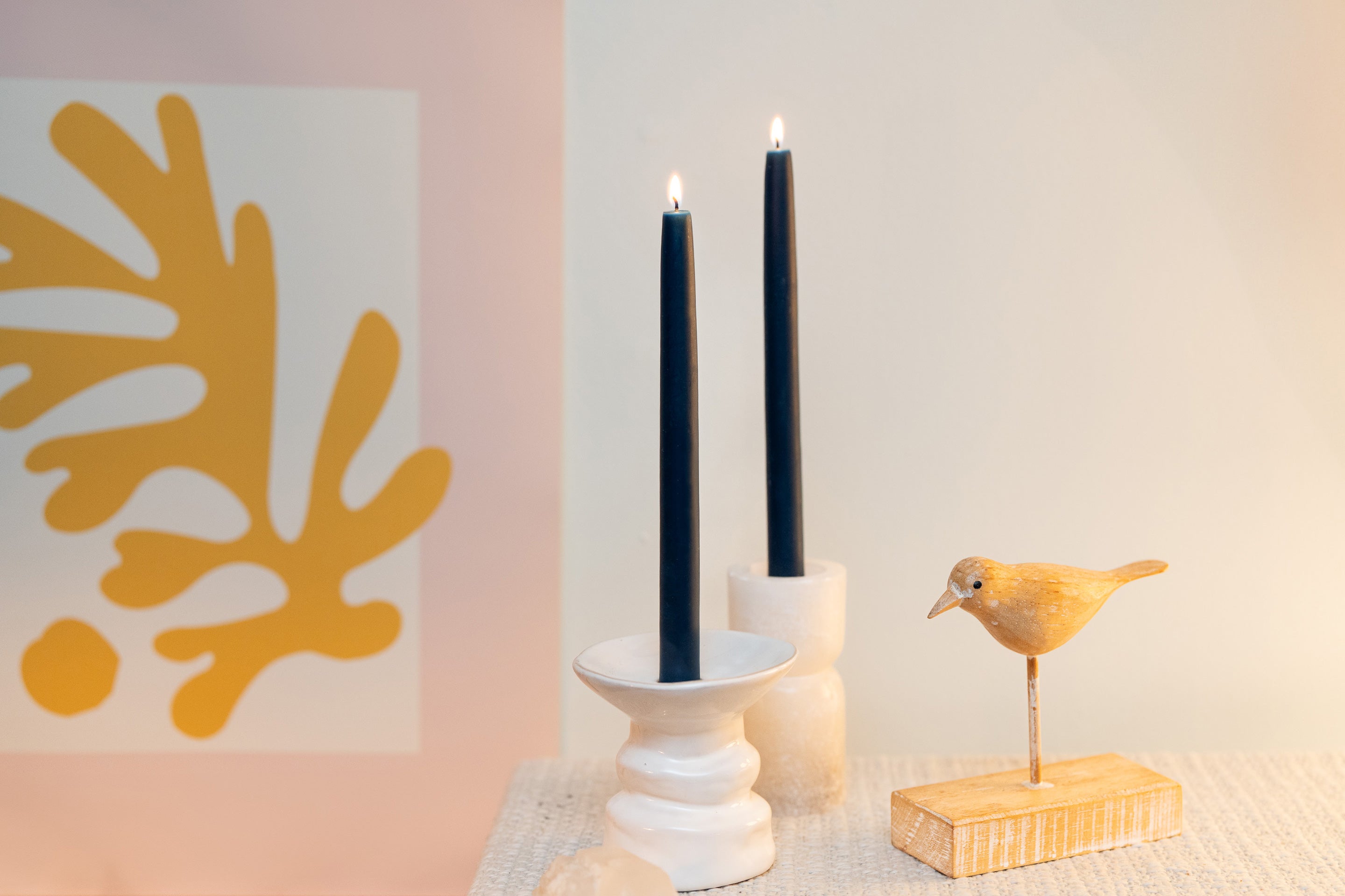Decorative setup with Sunbeam Candles 10" Dipped Beeswax Midnight Black taper candles in a white holder, a small yellow bird figurine, and abstract art on a light background.