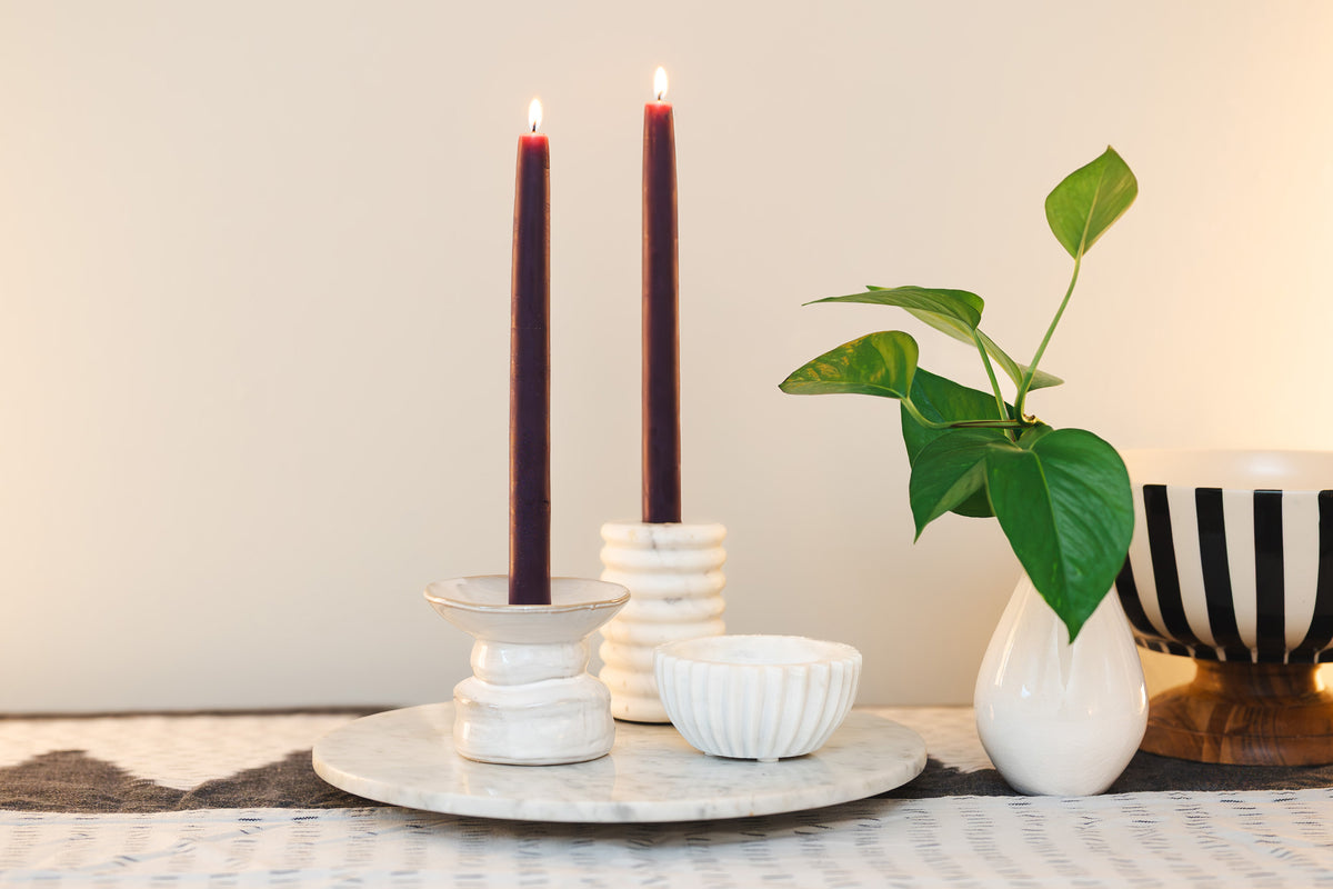 Two Sunbeam Candles 10" Dipped Beeswax Crimson Tapers lit in candle holders on a table with a white wall background and a plant next to them.