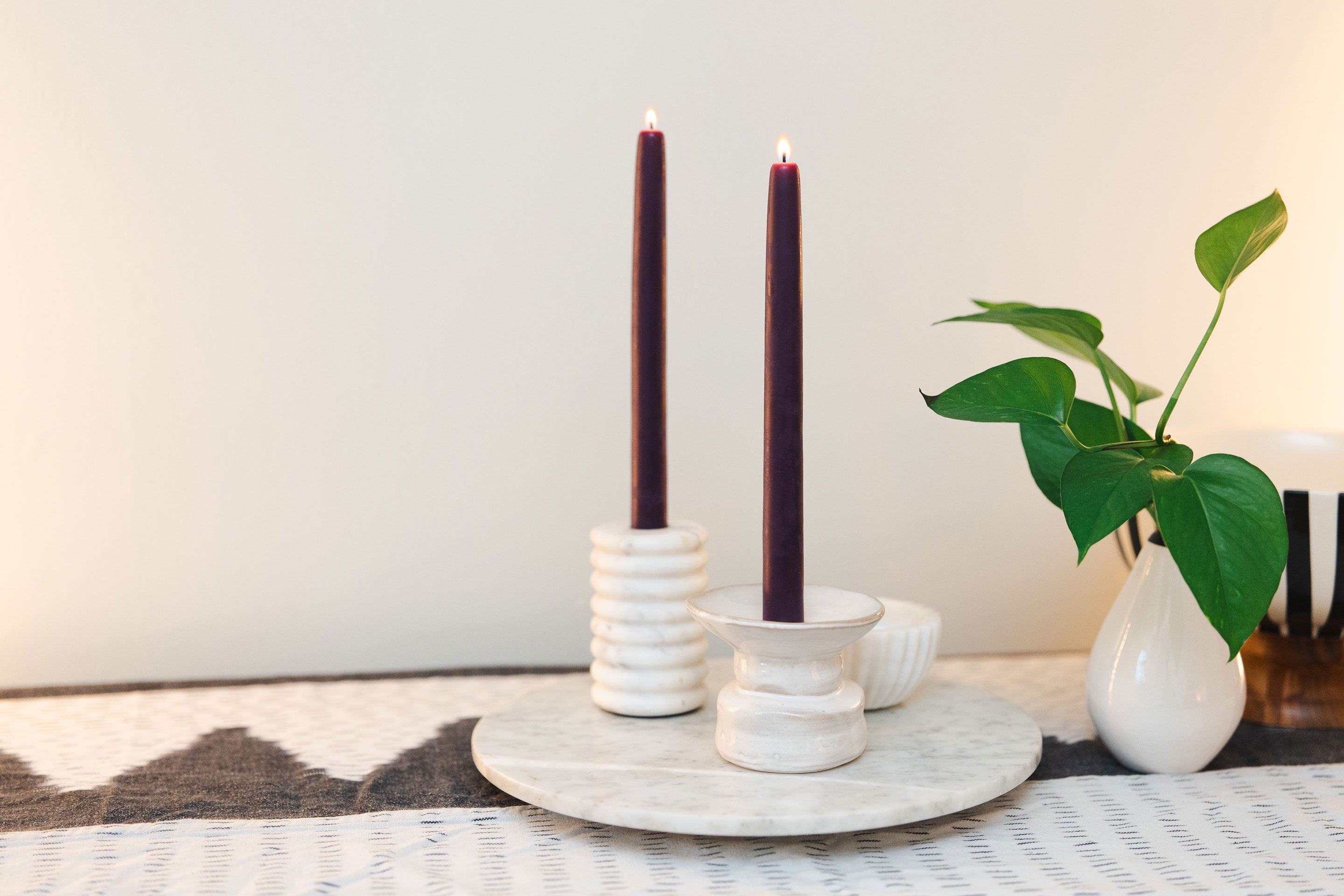 Two Sunbeam Candles 10" Dipped Beeswax Plum Tapers lit in candle holders with a white wall background and a plant next to them.