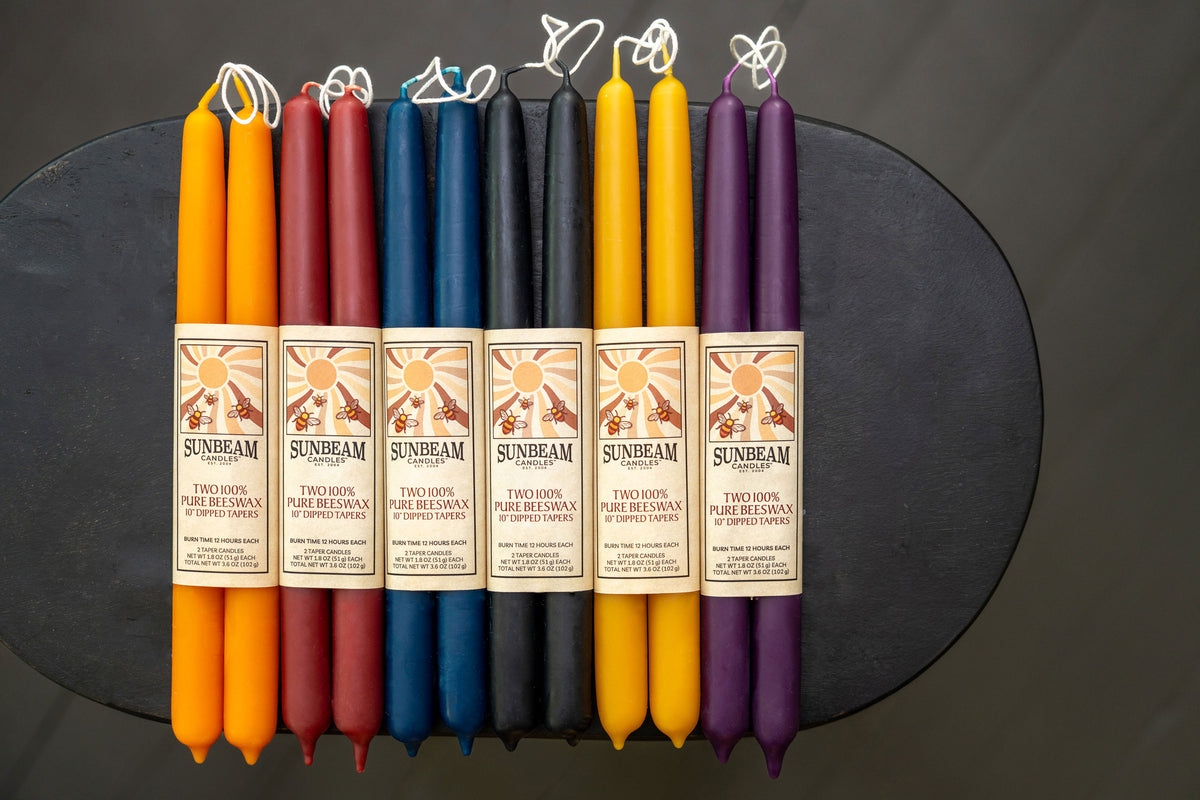 Sunbeam Candles 10" dipped beeswax tapers with wrapped labels in various colors on a black surface - Apricot, Crimson, Indigo, Midnight Black, Natural Gold & Plum