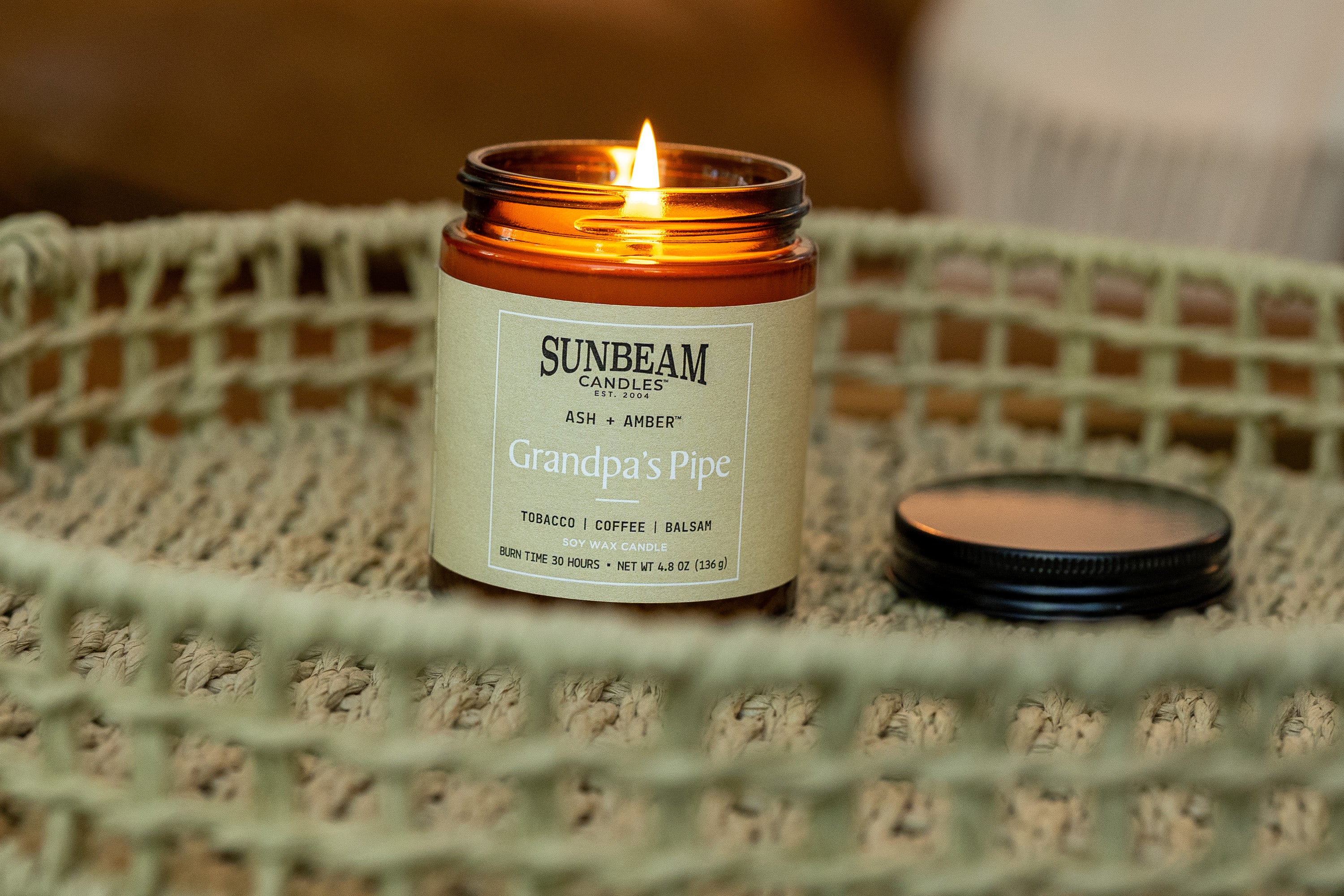 Sunbeam Candles Ash + Amber Grandpa's Pipe scented lit soy wax jar candle on wicker basket with lid next to it