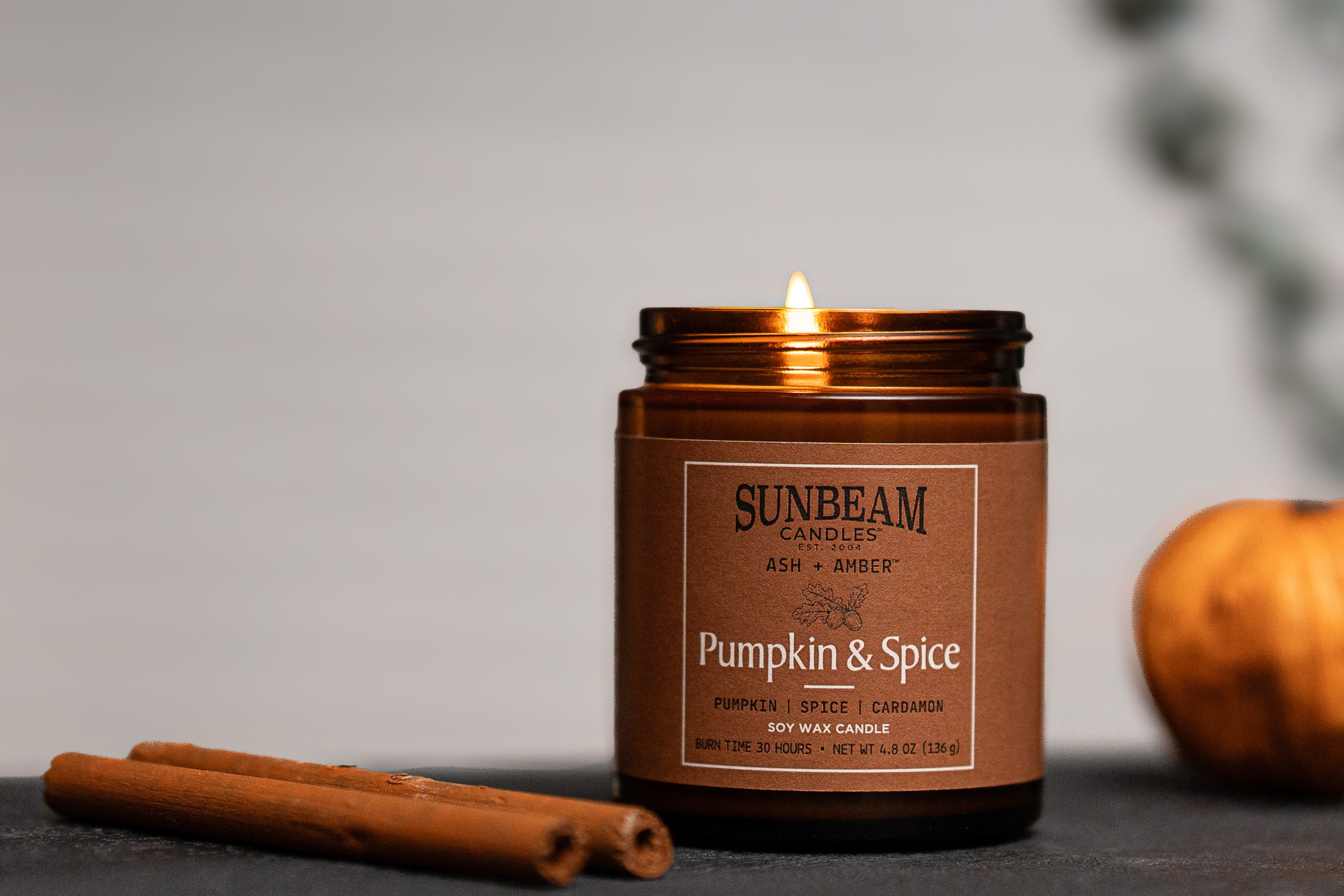 Sunbeam Candles Ash + Amber Fall Scent Pumpkin & Spice soy wax jar candles on a black table with a cinnamon stick next to it and a blurred background