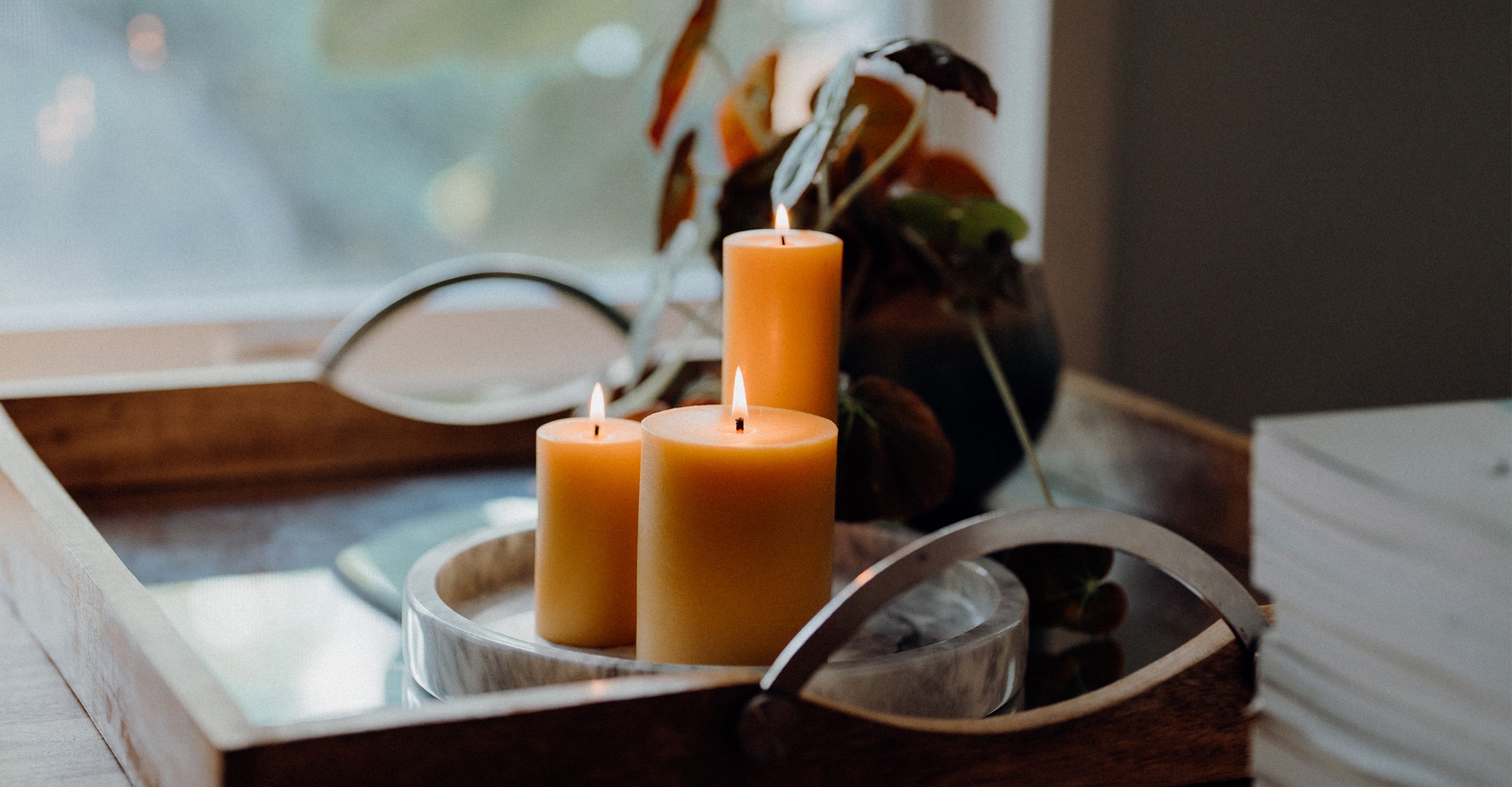 Three lit cream-colored pillar candles arranged in a round tray on a wooden surface, with a blurred interior background and dried botanicals behind them.