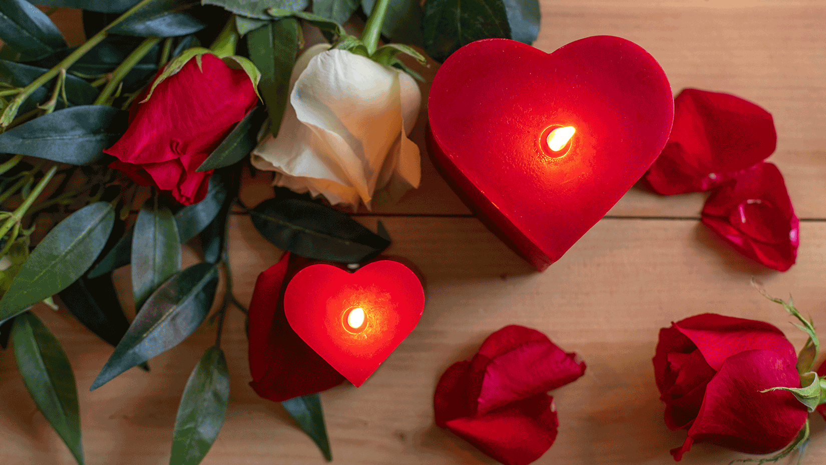 Sunbeam Candles Pure Beeswax Red Large & Small Heart Candles lit with flowers around them on a wood surface.