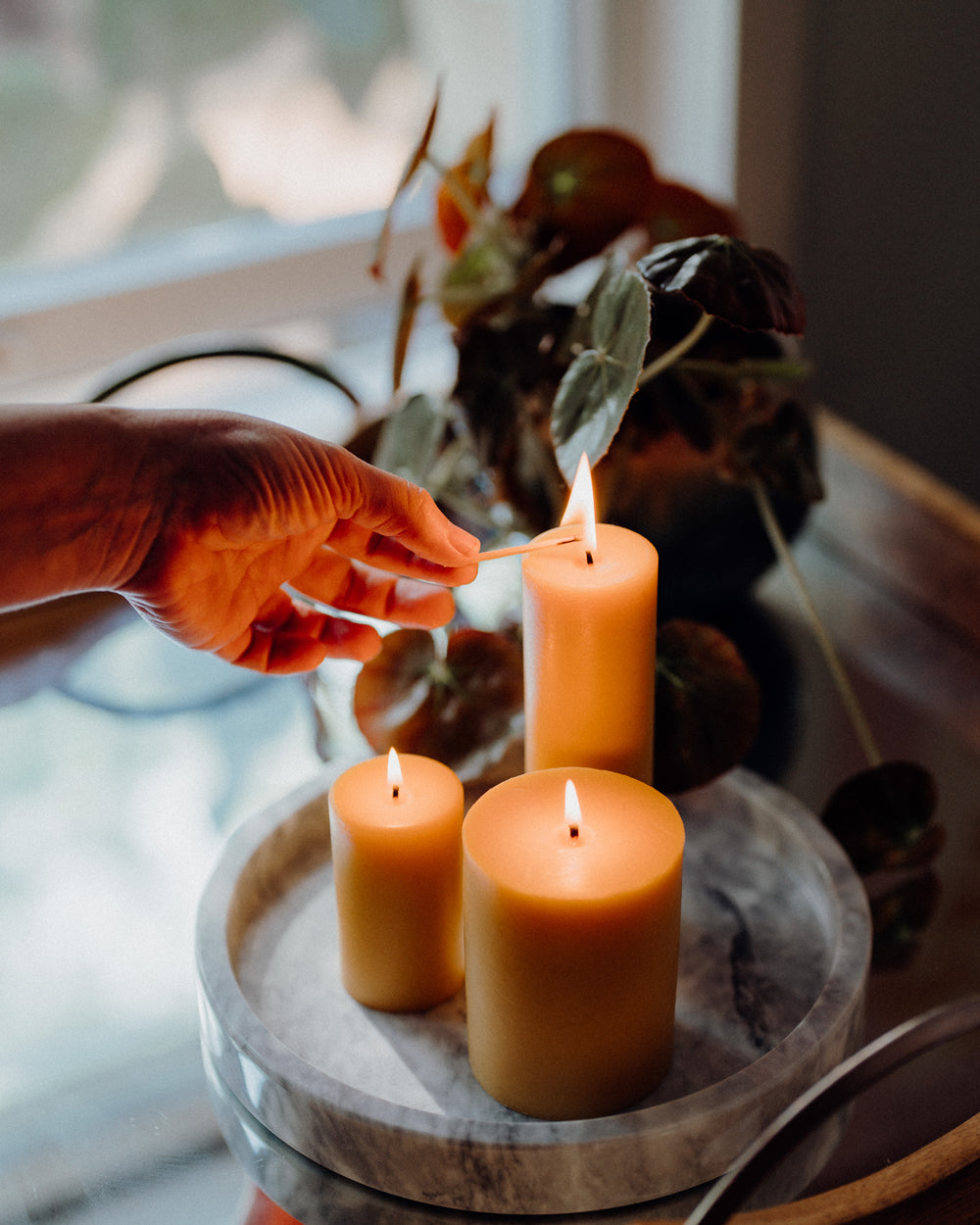Hand lighting three Sunbeam Candles Smooth Pillar candles on a wooden tray with a blurred background