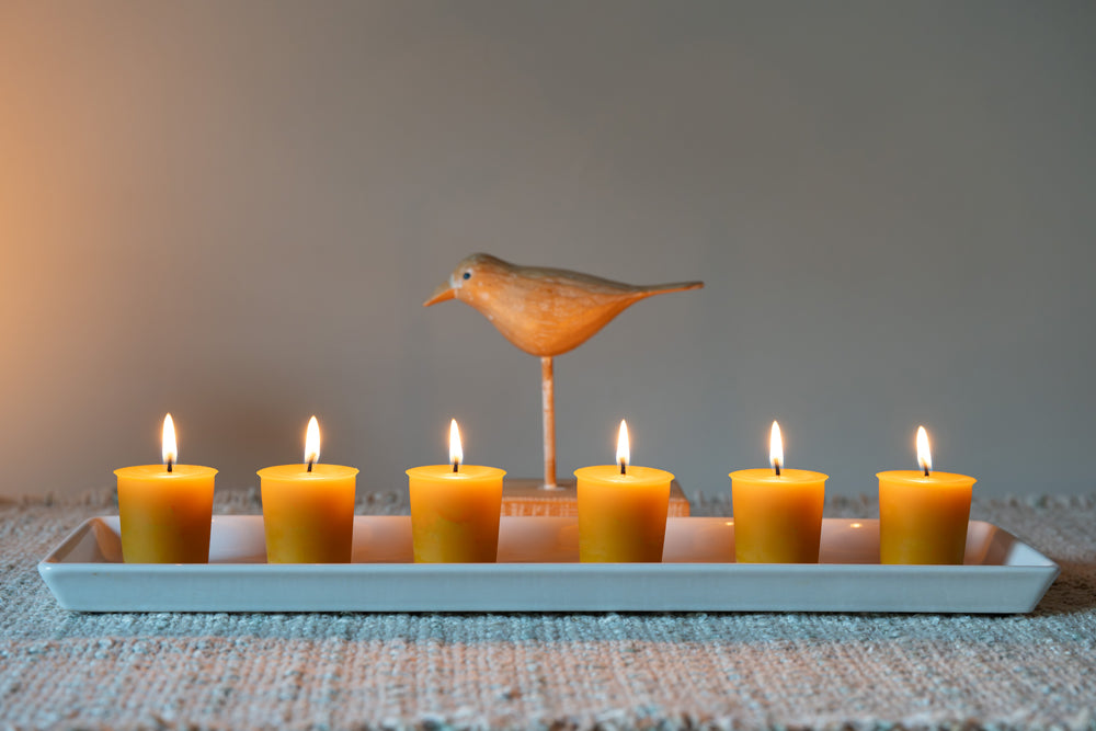 Row of lit candles on a white tray with a decorative bird figurine against a neutral background