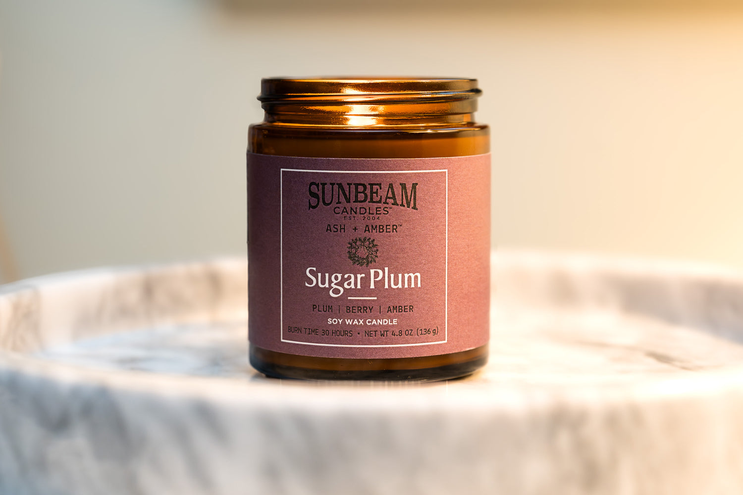 Sunbeam Candles Ash + Amber Fall Scent Sugar Plum soy wax jar candle on a plate with a blurred grey background