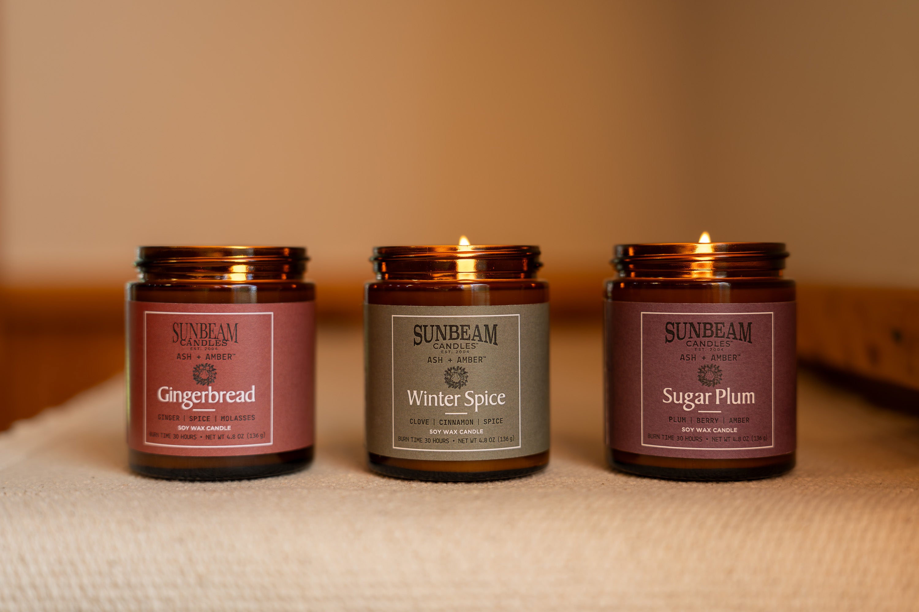 Sunbeam Candles Ash + Amber Winter Scent Soy Wax jar candles on a carpet with a yellowish-brown wall behind them