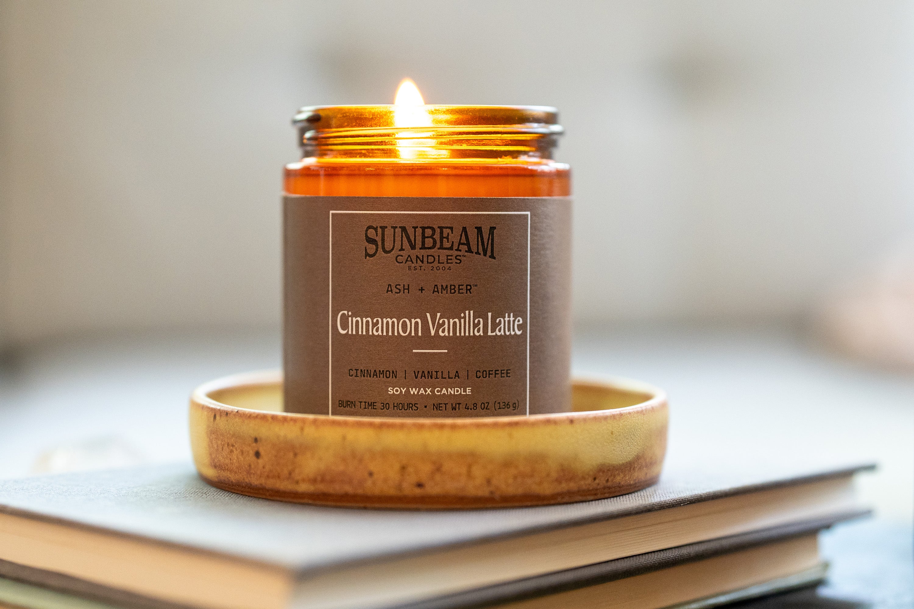 Sunbeam Candles Ash + Amber Everyday Scent Cinnamon Vanilla Latte soy wax jar candle in a candle holder sitting on a stack of books with a blurred background
