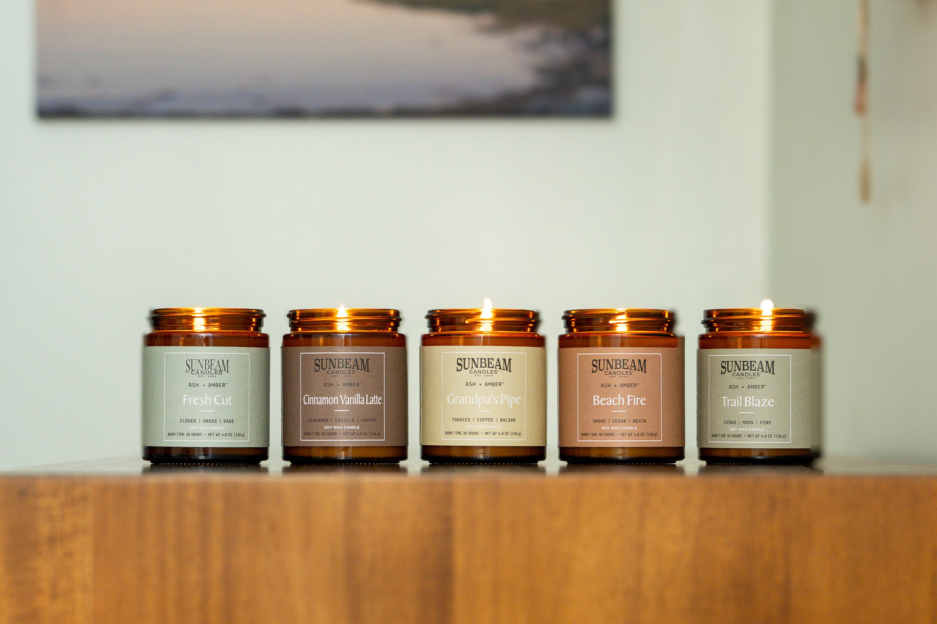 Sunbeam Candles Everyday Ash + Amber Soy Wax jar candles on a wood table with a white wall with a painting hanging on it