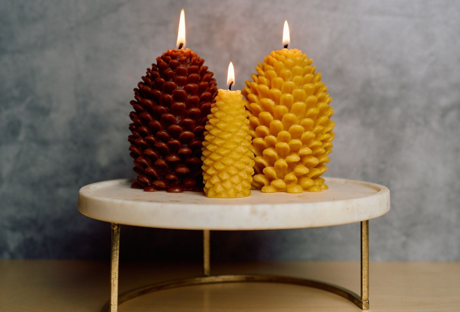 Three pine cone candles lit and sitting on a ceramic tray. Two large one in brown and one natural, one small in undyed natural wax.